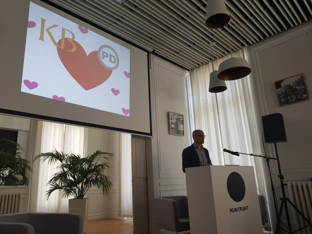 Mann an einem Rednerpult stehend und in ein Mikrofon sprechend, mit einem projizierten Bildschirm dahinter, umgeben von Stühlen, einer Pflanze, Fotorahmen, Vorh├Ąngen und Deckenleuchten.