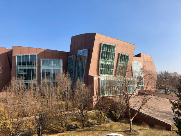 Großes Gebäude mit vielen Fenstern, umgeben von Bäumen, Bänken, Gras und einem Laternenmast, mit anderen Gebäuden und einem klaren blauen Himmel im Hintergrund.