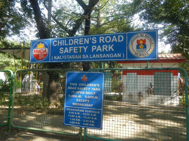 Ein von einem Metallzaun umgebener Kinderverkehrssicherheitspark mit zwei blauen Informationsschildern, einem kleinen Haus mit Dach, einigen Pflanzen, einer Gruppe von Bäumen und einem bewölkten Himmel.