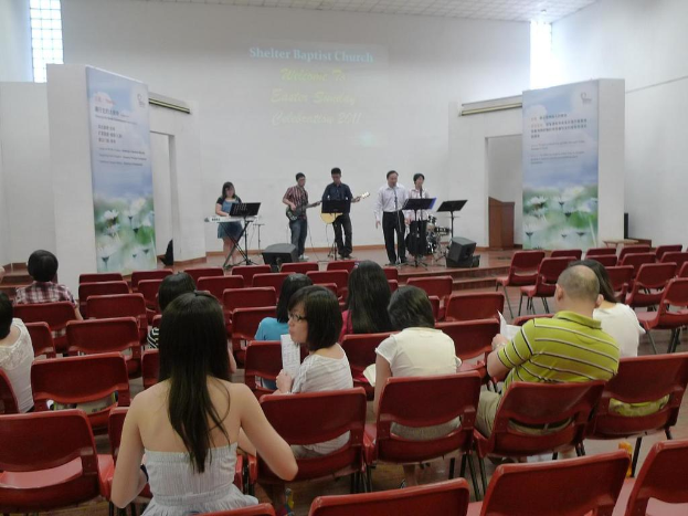 Eine Bühne mit zwei Gitarristen in der Mitte und anderen Musikern in den Ecken, die singen und Instrumente spielen, sowie Schilder neben der Bühne und ein Publikum davor mit Papieren und Büchern.