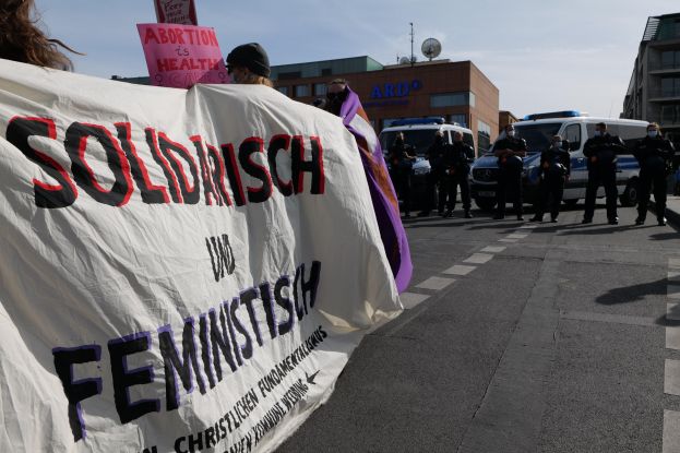 Eine Gruppe von Menschen marschiert die Straße entlang, hält ein Banner mit der Aufschrift "Solidarität und Feminismus" in der Hand, mit parkenden Fahrzeugen am Straßenrand und Gebäuden im Hintergrund unter einem klaren blauen Himmel.