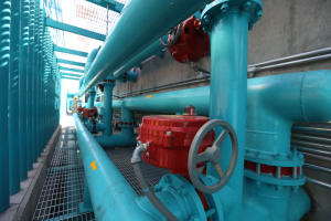 Eine große Gruppe von blauen Rohren und Ventilen in einem Gebäude, mit einem prominenten roten Ventil in der Mitte, Metallstangen links und einer Wand im Hintergrund.