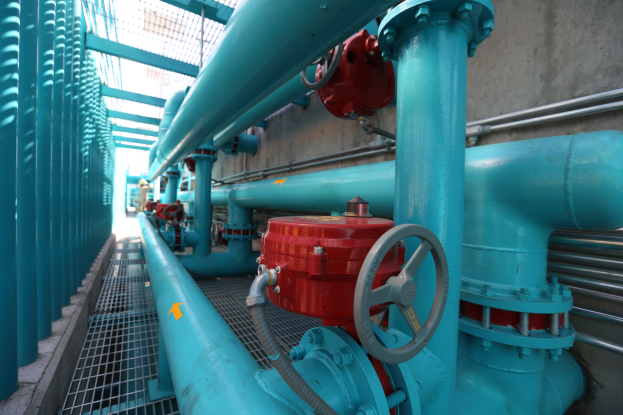 Eine große Gruppe von blauen Rohren und Ventilen in einem Gebäude, mit einem prominenten roten Ventil in der Mitte, Metallstangen links und einer Wand im Hintergrund.