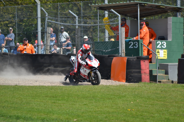 Ein Motorradfahrer in Helm und Handschuhen fährt über ein grasbewachsenes Feld mit Zuschauern in Mützen und Schutzbrillen, einem eingezäunten Bereich, Treppen mit Geländern und einem nummerierten Schuppen im Hintergrund.