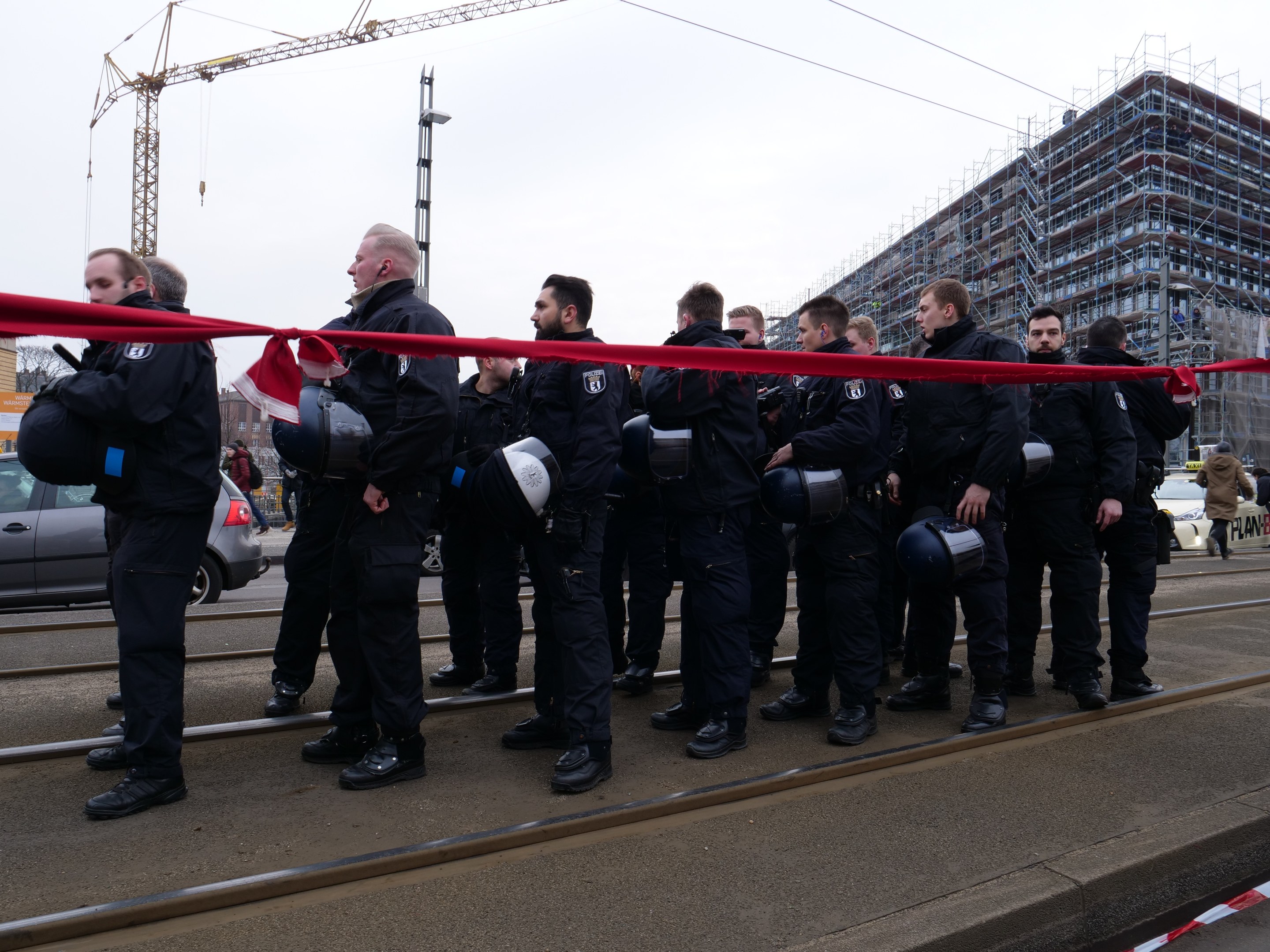 Gruppe von Polizisten in schwarzen Uniformen und Helmen auf einem Bahnsteig mit einem roten Band davor, Fahrzeuge und Menschen im Hintergrund unter einem klaren blauen Himmel.