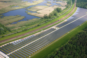 Eine Luftaufnahme einer Solarpark mit zahlreichen Solarpanelen in einem Feld umgeben von Bäumen, Gras und Wasser, mit einem Zug, der auf einem Bahnsteig in der Nähe fährt.