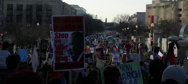 Eine Gruppe von Menschen, die auf der Straße protestieren und Plakate halten, mit Gebäuden, Bäumen und Laternen im Hintergrund unter einem klaren Himmel.
