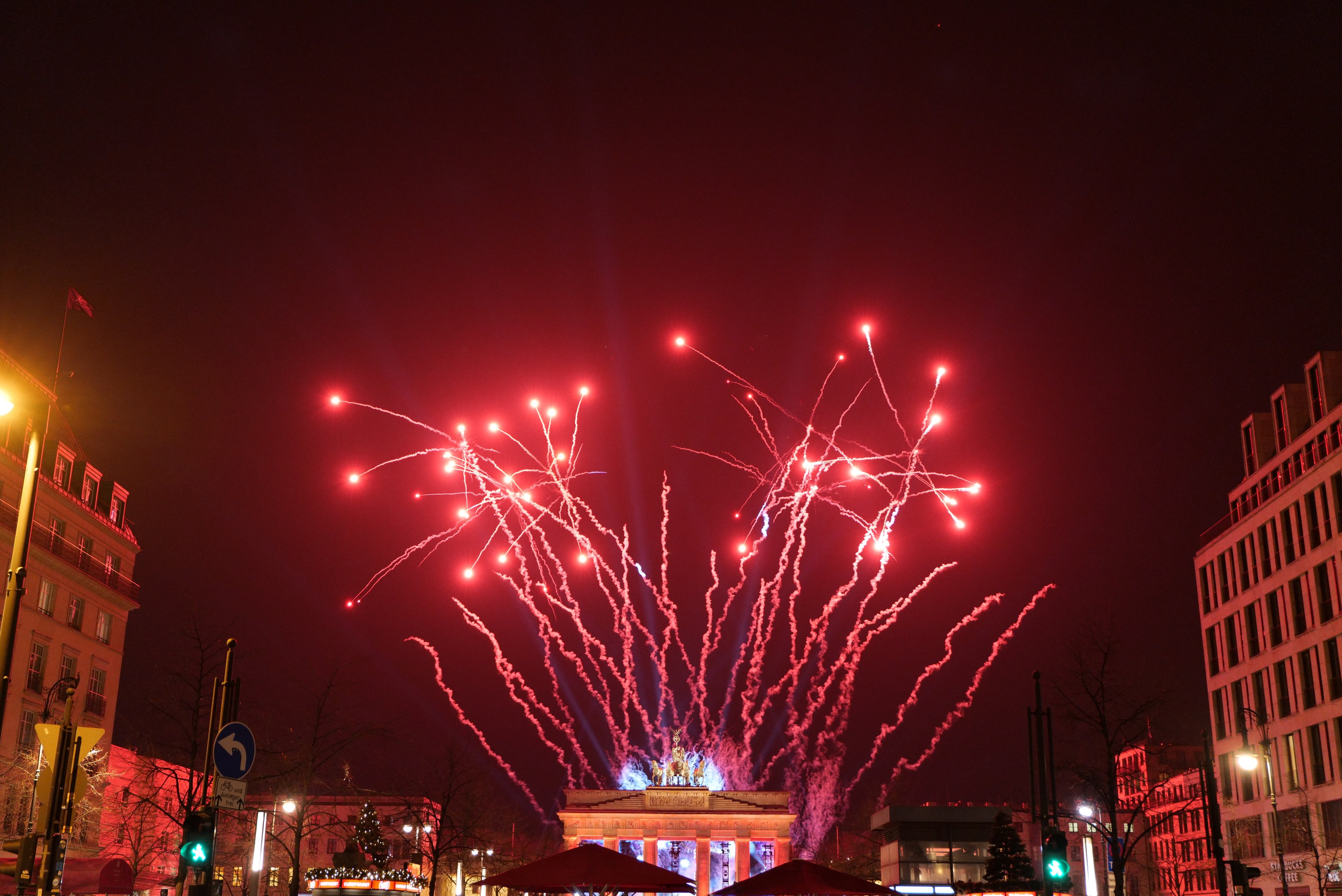 Eine nächtliche Stadtstraße an Silvester in Berlin mit Gebäuden, Bäumen, Laternen, Ampeln, Schildern, Zelten, Menschen und einem prächtigen Feuerwerk am Himmel.