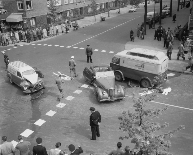 Schwarz-weiß-Szene eines Autounfalls an einer Straßenkreuzung mit mehreren Fahrzeugen, darunter ein Lieferwagen, und einer Gruppe von Zuschauern; Gebäude, Bäume, Strommasten und Texttafeln sind im Hintergrund zu sehen.