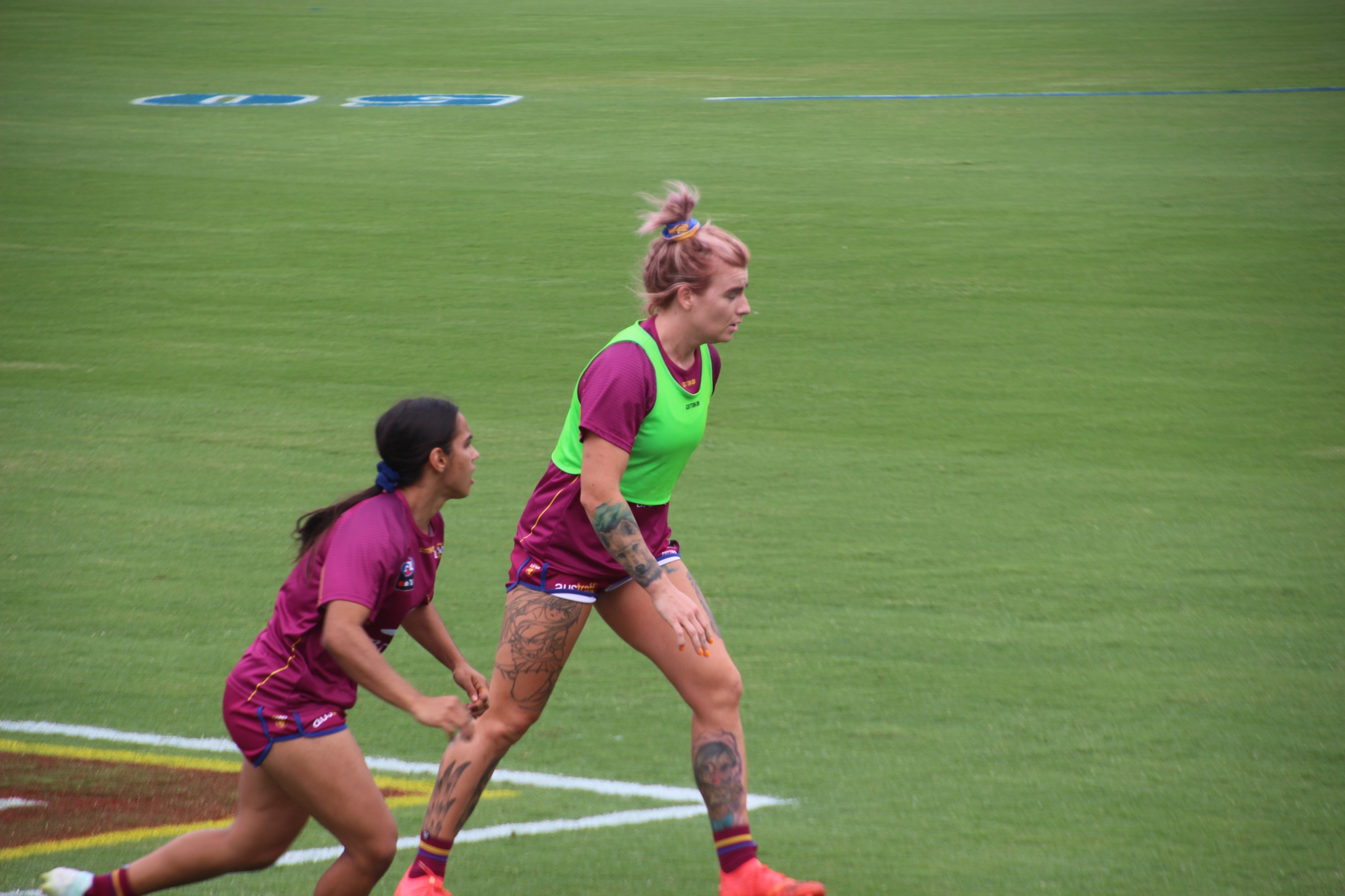 Zwei Frauen in Sportkleidung, eine in einem grünen T-Shirt und Shorts und die andere in einem pinken Kleid, spielen Fußball auf einem Feld und zeigen sichtbare Tattoos an Händen und Füßen.