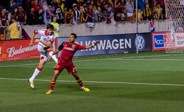 In der Mitte spielen Personen auf einem grünen Feld. Im Hintergrund hält jemand in einem grünen T-Shirt einen Ball, während andere zuschauen. Banner mit Text sind sichtbar, und ein weißes Netz befindet sich rechts vor einem Banner.