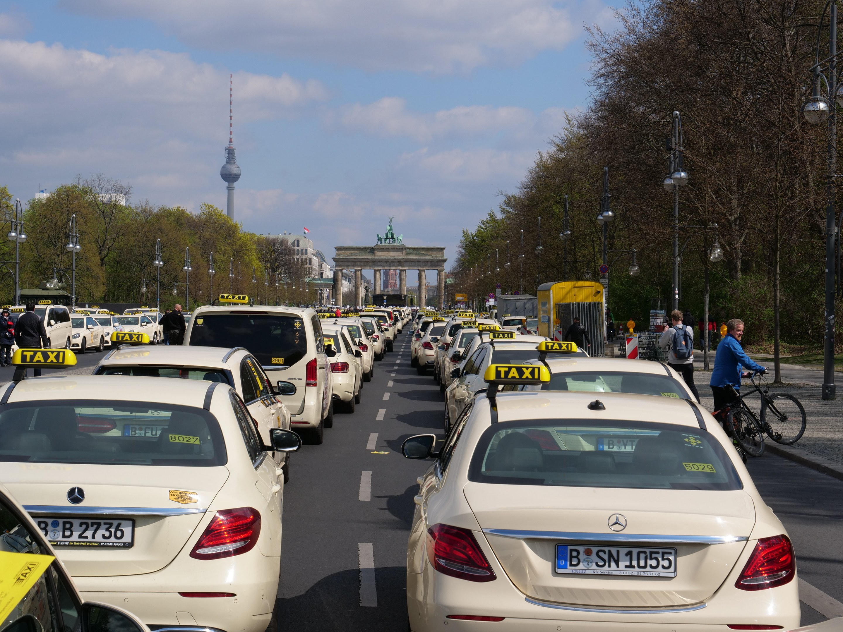 Eine lange Reihe von Taxis entlang einer belebten Straße in Berlin, Deutschland, mit Fahrradfahrern und Fußgängern auf dem Gehweg, gesäumt von Bäumen und Laternenmasten und Gebäuden, einem Bogen und einem Turm im Hintergrund unter einem bewölkten Himmel.