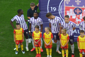 Eine Gruppe junger Menschen auf einem grünen Fußballfeld mit einem "Ligue 1"-Plakat im Hintergrund, einige tragen Trikots und andere Shorts, unter einem hellblauen Himmel.