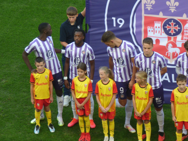 Eine Gruppe junger Menschen auf einem grünen Fußballfeld mit einem "Ligue 1"-Plakat im Hintergrund, einige tragen Trikots und andere Shorts, unter einem hellblauen Himmel.