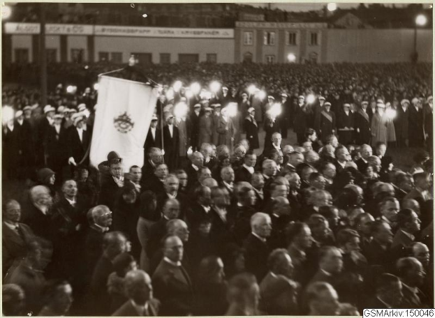 Schwarzes und weißes Foto einer großen Menschenmenge vor einem Gebäude, mit einer Person, die eine Fahne hält, Stangen und Lichter im Hintergrund und Text unten mit der Aufschrift "Gustav Klimts Rede an der Berliner Mauer."