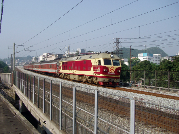 Ein roter und gelber Zug fährt auf Schienen neben einer Brücke, einem Geländer, Strommasten mit Drähten, Bäumen, Gebäuden und einem klaren blauen Himmel.