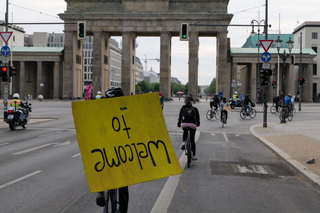 Eine Gruppe von Menschen fährt auf Fahrrädern eine Straße in Berlin, Deutschland, vor dem Brandenburger Tor entlang, mit Helmen, einer Person mit einer gelben Schilder, Laternen, Verkehrszeichen, Gebäuden, Bäumen und einem klaren blauen Himmel im Hintergrund.