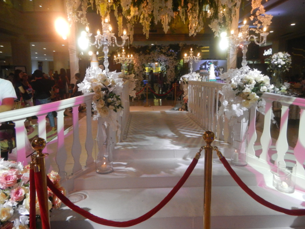 Eine Hochzeitszeremonie in einem Einkaufszentrum mit weißem Teppich, dekorativen Geländern, Blumenschmuck, Kronleuchtern und Deckenbeleuchtung, bei der eine Menschenmenge dabei ist, einschließlich Personen mit Taschen.