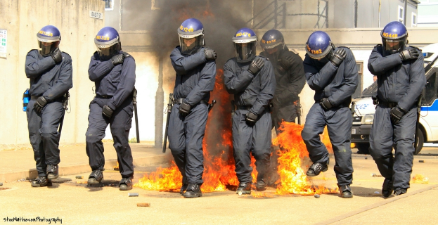 Menschen in Helmen stehen vor einem Feuer, mit Gebäuden, einem Fahrzeug und verschiedenen Gegenständen im Hintergrund; ein Plakat und eine Tafel sind an einer Wand auf der linken Seite befestigt, und Text ist am unteren Rand des Bildes sichtbar.
