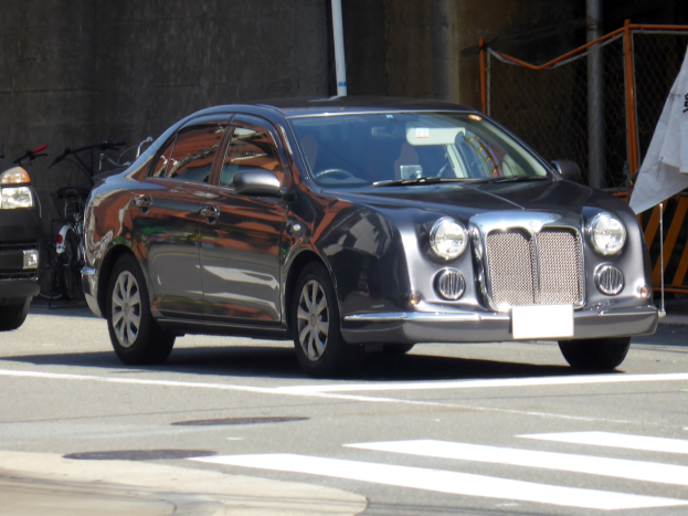 Ein silbernes Auto, das auf der Straße neben einem Fahrrad geparkt ist, mit einer Wand im Hintergrund und einem Maschendrahtzaun auf der rechten Seite.