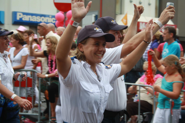 Eine Gruppe von Menschen, einige mit Mützen und Dosen haltend, steht und sitzt vor einer Menge, mit Geländern, Ballons und einem Gebäude mit Schildern im Hintergrund.