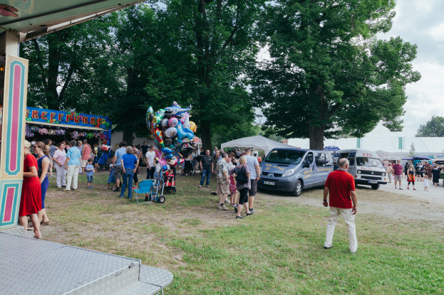 Eine Gruppe von Menschen, einige mit Mützen, steht vor einer Karussellfahrt, umgeben von Gras, Fahrzeugen, Buden, Luftballons, Bäumen und einem bewölkten Himmel.
