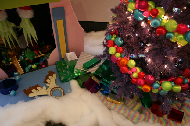 Drinnen steht ein geschmückter Weihnachtsbaum mit Lichterketten und Schmuck rechts, umgeben von Geschenkboxen und Dekorationsgegenständen, mit einer Hütte und Baumwolle im Hintergrund.