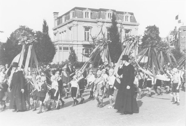 Schwarzes und weißes Bild einer Gruppe von Menschen, die eine Straße entlanggehen und Fahnen halten, mit Hüten, Maibaum, Bäumen, Gebäuden und Himmel.