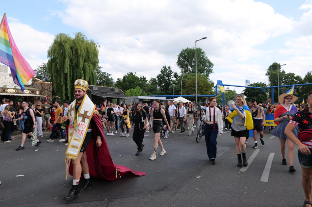 Eine Gruppe von Menschen bei der 2018er Gay Pride Parade mit einer Regenbogenfahne und Musikinstrumenten, einige tragen Mützen, vor einem Hintergrund aus Laternenmasten, Bäumen, Schuppen und einem bewölkten Himmel.