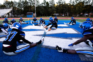 Gruppe junger Männer in Sportkleidung, Helmen und Schuhen sitzend auf einem Fußballfeld mit Bäumen, Pfosten, Gebäuden und einem klaren blauen Himmel im Hintergrund, neben einer Treppe mit Geländern.