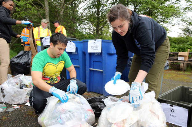 Zwei Personen in Handschuhen sammeln Müll auf Tellern in einem Park, umgeben von weggeworfenen Flaschen und Plastik, mit einem Mülleimer und einer Bank rechts und Bäumen und Himmel im Hintergrund.