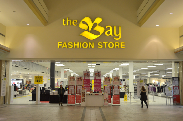 Exterior view of a Bay fashion store in a mall with shoppers, wall text boards, pillars, ceiling lights, and background displays.