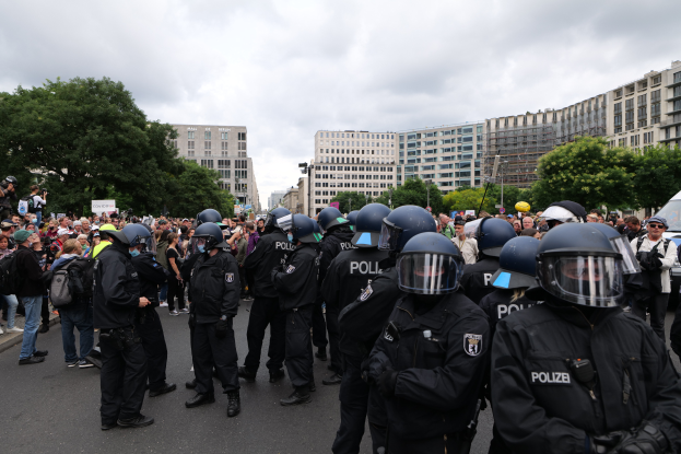 Eine große Gruppe von Polizisten steht vor einer Menschenmenge auf einer Straße, die von Bäumen und Gebäuden mit Fenstern umgeben ist, mit einem bewölkten Himmel im Hintergrund, wobei einige Menschen in der Menge Kameras halten, was auf eine Demonstration in Berlin, Deutschland, hindeutet.