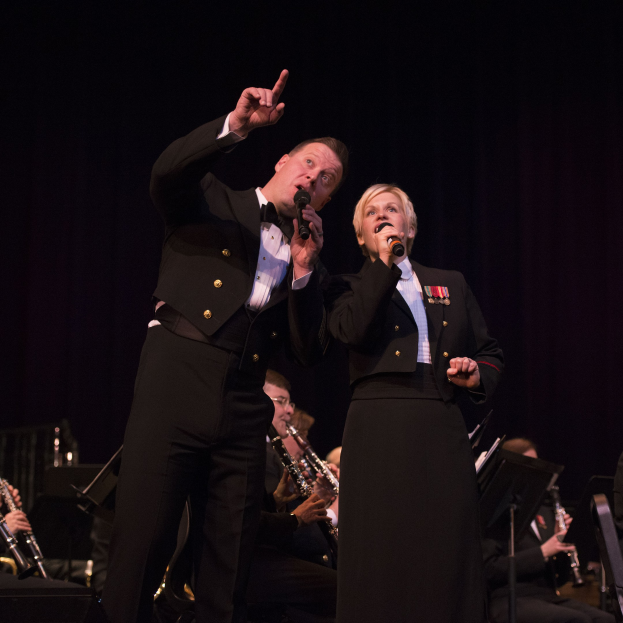Ein Mann und eine Frau in Smokings stehen auf einer Bühne und halten Mikrofone, mit einem sitzenden Orchester dahinter vor einem dunklen Hintergrund.