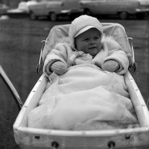 Ein Schwarz-Weiß-Foto eines lachenden Babys in einem Kinderwagen, das eine weiße Kleidung und eine Kappe trägt, mit Fahrzeugen im Hintergrund.