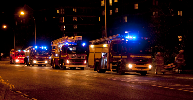 Feuerwehrlöschfahrzeuge sind nachts auf der Straße, mit Gebäuden und Laternen im Hintergrund und zwei Personen auf der rechten Seite.