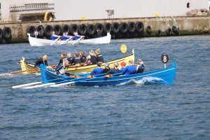 Eine Gruppe von Menschen in einem blauen und gelben Boot auf dem Wasser, die Paddel halten, mit einer Wand mit Reifen und einem Gebäude im Hintergrund, was auf eine Regatta hindeutet.