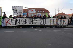Protestierende mit einem Banner mit der Aufschrift 'Kein Grund zu Feiern' gegen deutsche Sparmaßnahmen vor einem Gebäude mit Bäumen und einem klaren Himmel.