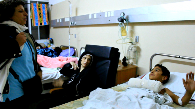 Ein Krankenhauszimmer mit mehreren Betten, die von Patienten belegt sind, Infusionsflaschen auf Ständern zwischen den Betten und eine Frau in der Nähe eines Patienten mit einem Beutel.