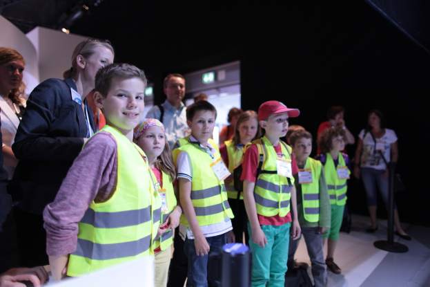 Eine Gruppe von Kindern in Sicherheitswesten und Namensschildern, mit einem Jungen, der links lächelt, auf einem Boden mit einem Pfahl rechts und einigen Menschen im Hintergrund.