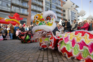 Ein lebendiges chinesisches Neujahrsfest in Amsterdam mit einem Löwen tanzen im Vordergrund und einer Menge Menschen drumherum, einige halten Kameras, vor einem Hintergrund aus Gebäuden, Laternenmasten und einem klaren blauen Himmel.