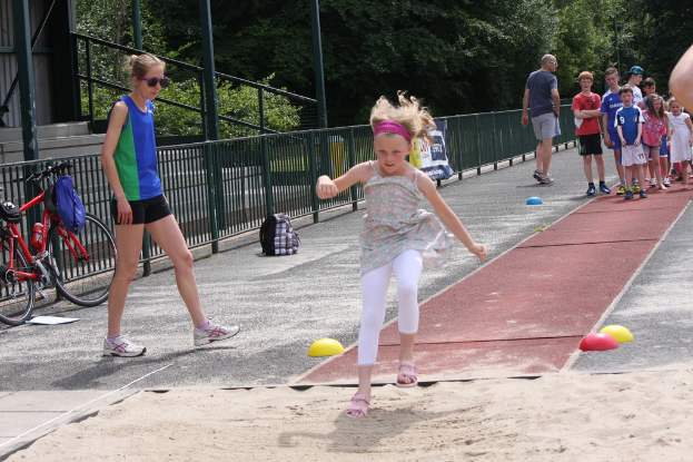 Ein Mödchen steht im Mittelpunkt des Bildes, umgeben von spielenden Menschen, mit einem Fahrrad, einer Begrenzung links und Bäumen und Treppen im Hintergrund.