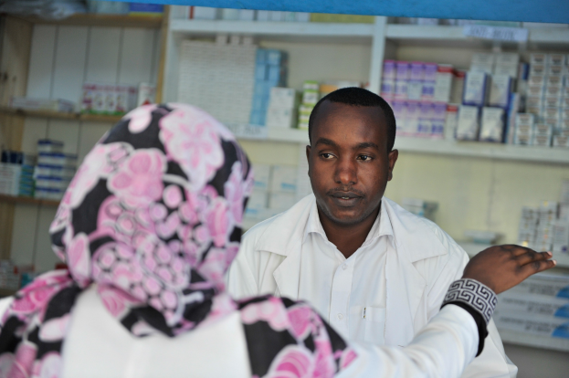 Apothekerin in weißem Kittel, die zu einer sitzenden Frau in einer Apotheke spricht, mit Regalen voller Schachteln und Gegenständen im Hintergrund.