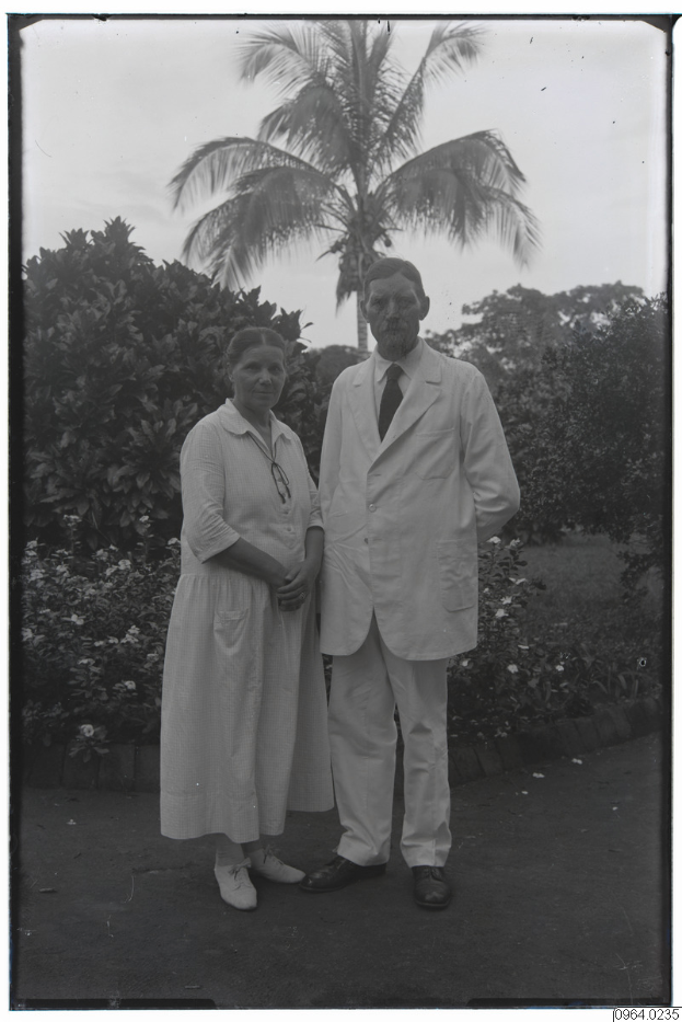 Ein altes Schwarz-Weiß-Foto eines Mannes in einem Anzug und einer Frau, die nebeneinander vor einer Palme stehen, mit Bäumen und einem klaren Himmel im Hintergrund.