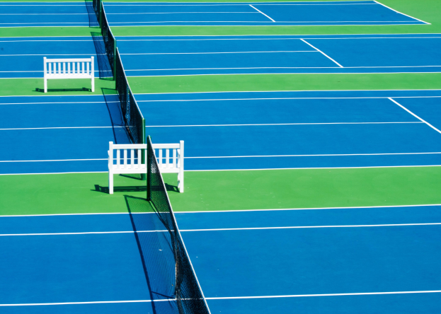 Ein blau-grüner Tennisplatz mit zwei weißen Bänken, einem Netz in der Mitte und umgeben von saftig grünem Gras.