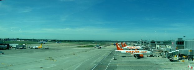 Eine Flughafen-Szene mit Flugzeugen, Gebäuden, Pfählen, Fahrzeugen, Bäumen und einem Himmel darüber sowie einer Straße darunter und einem Zaun im Vordergrund.