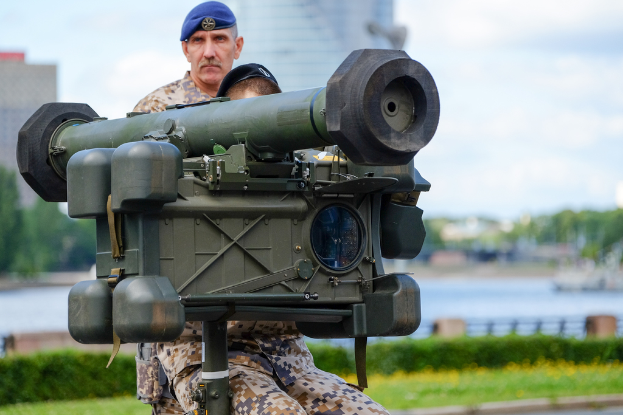 Zwei Personen in militärischer Uniform mit grünen Waffen vor einem See, mit Gebäuden, Bäumen und einem klaren Himmel im Hintergrund.