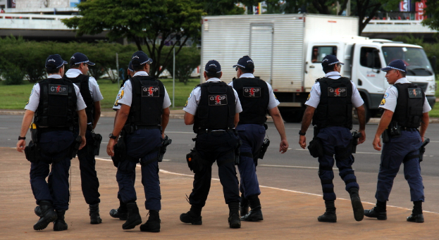 Eine Gruppe von Beamten in Uniform, einschließlich Mützen, Hemden, Schutzwesten, Hosen und Schuhen, geht auf einer Straße. Rechts daneben steht ein Lastwagen in der Nähe eines Zauns, mit Gebäuden, Bäumen, Pflanzen, Pfählen und Bannern im Hintergrund und einer Brücke oben rechts.