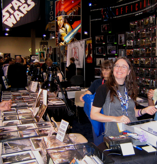 Eine Frau posiert an der Kasse in einem belebten Geschäft mit Bildern, Büchern und ausliegenden Plakaten, während Kunden im Hintergrund einkaufen.