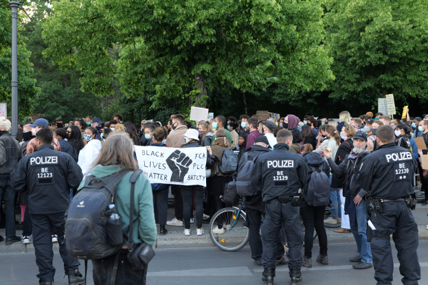Eine große Gruppe von Menschen nimmt an einer Black-Lives-Matter-Demonstration in Berlin teil, einige halten Schilder und tragen Kappen und Taschen, mit einem Fahrrad im Vordergrund und Bäumen und einem Pfahl im Hintergrund.